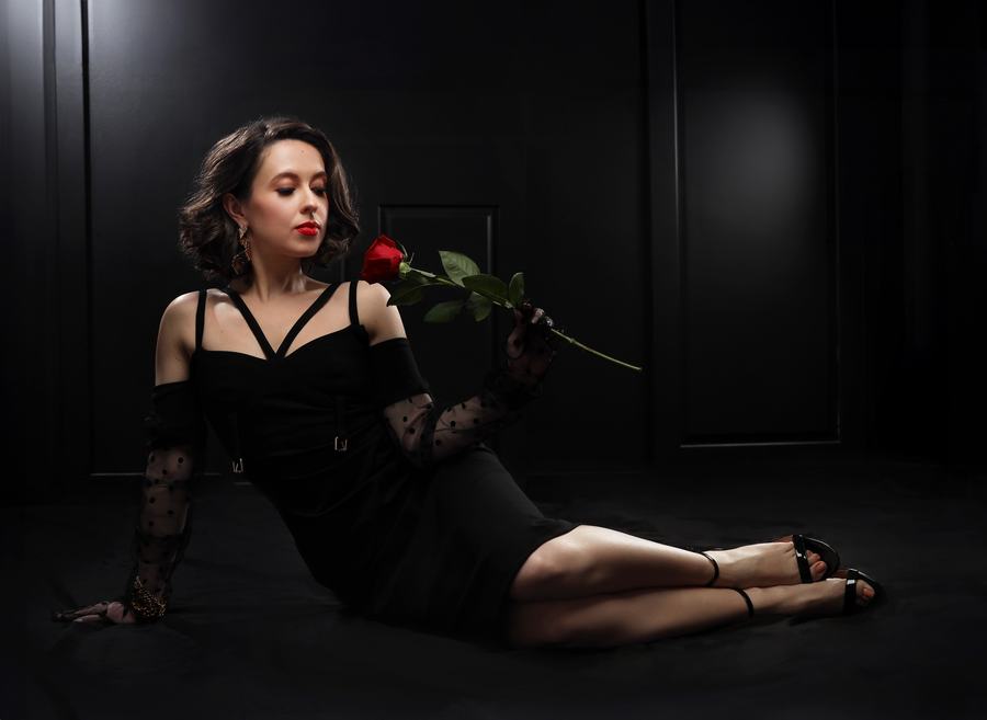 Reclining in black on a dark set, a red rose held to the lips