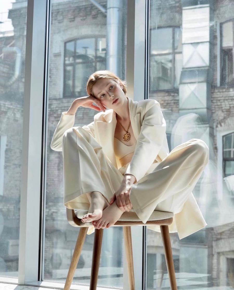 Cream tailoring, seated barefoot on a stool before tall windows