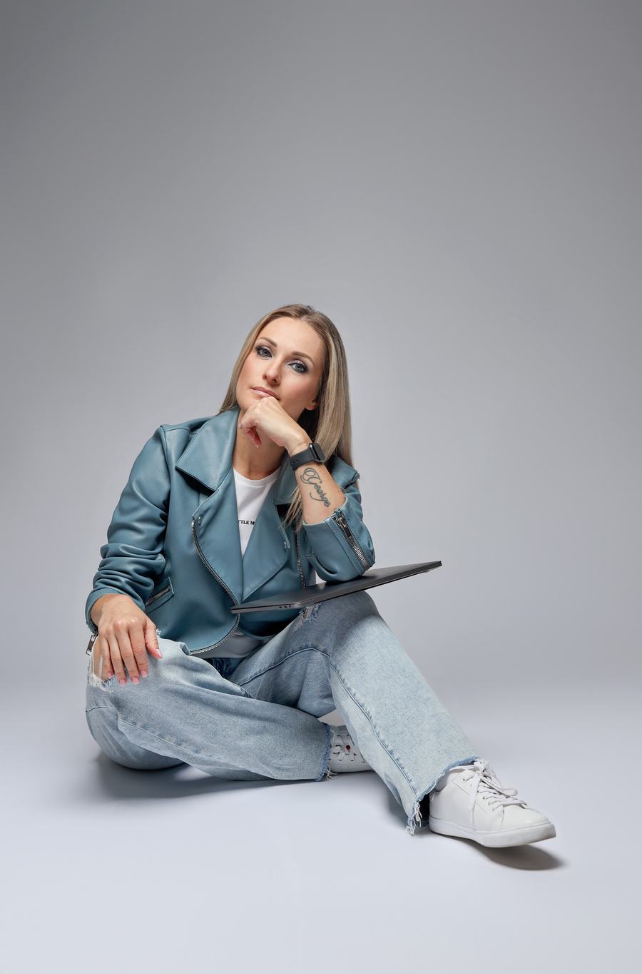 Seated on a grey seamless in a teal leather jacket, laptop in hand